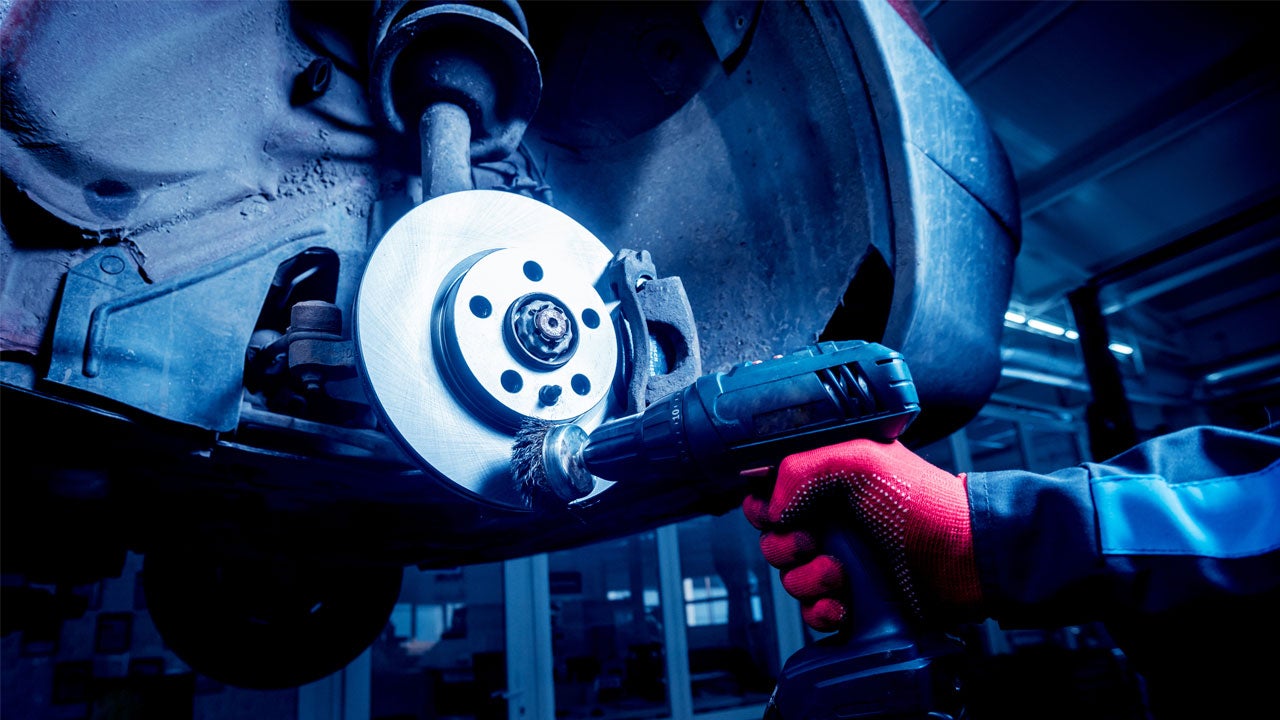 A mechanic cleaning a vehicle's brake rotor surface using a power drill with a wire wheel attachment.